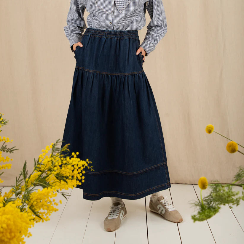 Woman wearing a blue blouse with a bow and a navy skirt, standing against a beige background with yellow flowers.