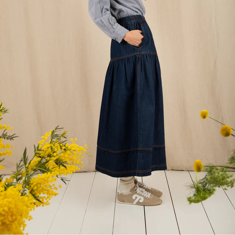 Woman in a denim skirt and gray blouse standing against a beige wall with yellow flowers on a white floor.