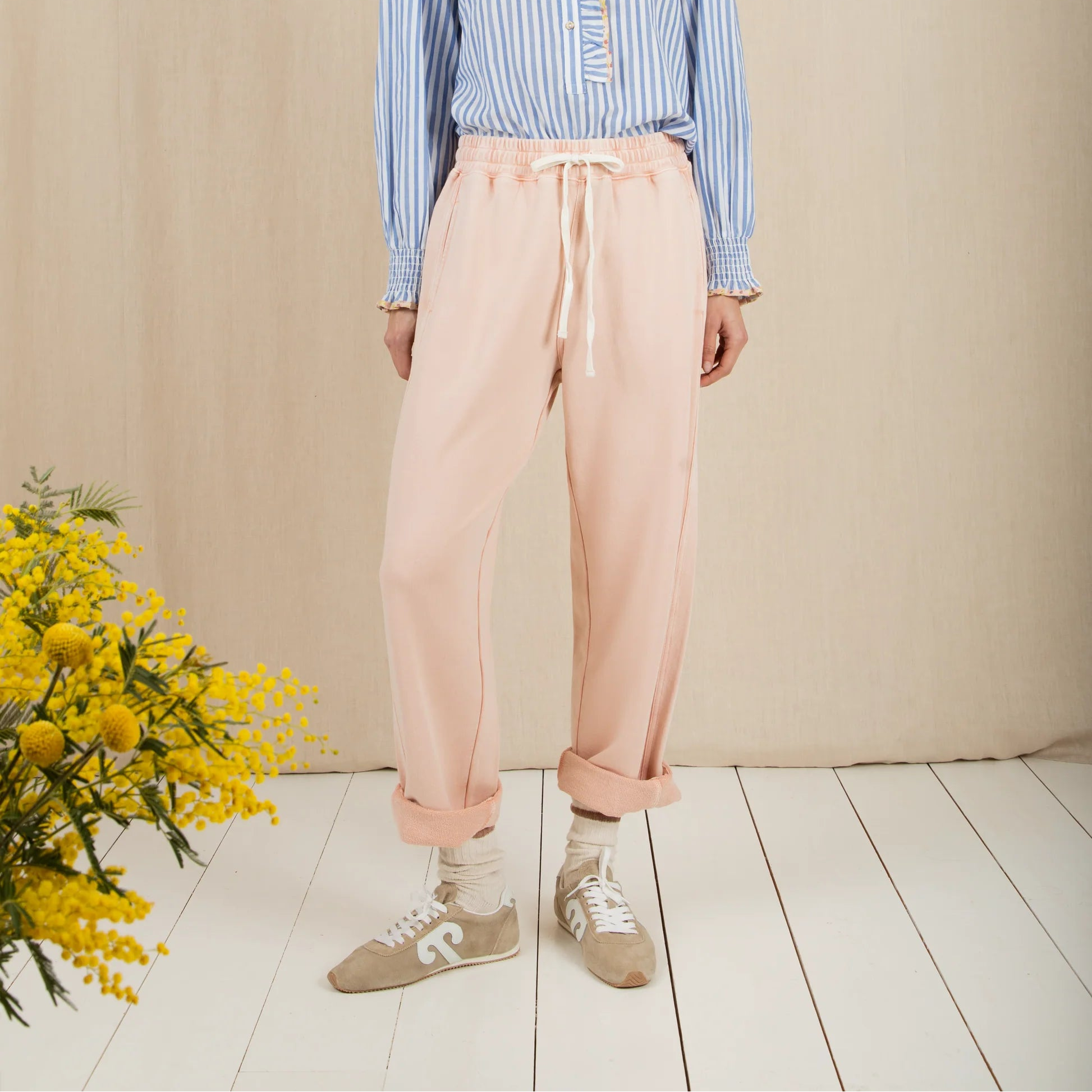 Woman wearing a blue striped shirt and pink pants standing against a beige wall with a bouquet of yellow flowers on the left.
