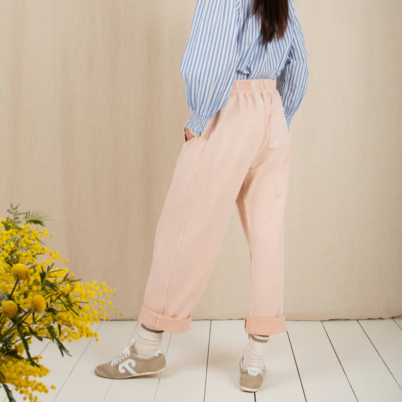 Woman wearing a blue striped shirt, pink pants, and a white cap standing against a beige wall with a plant on the left.