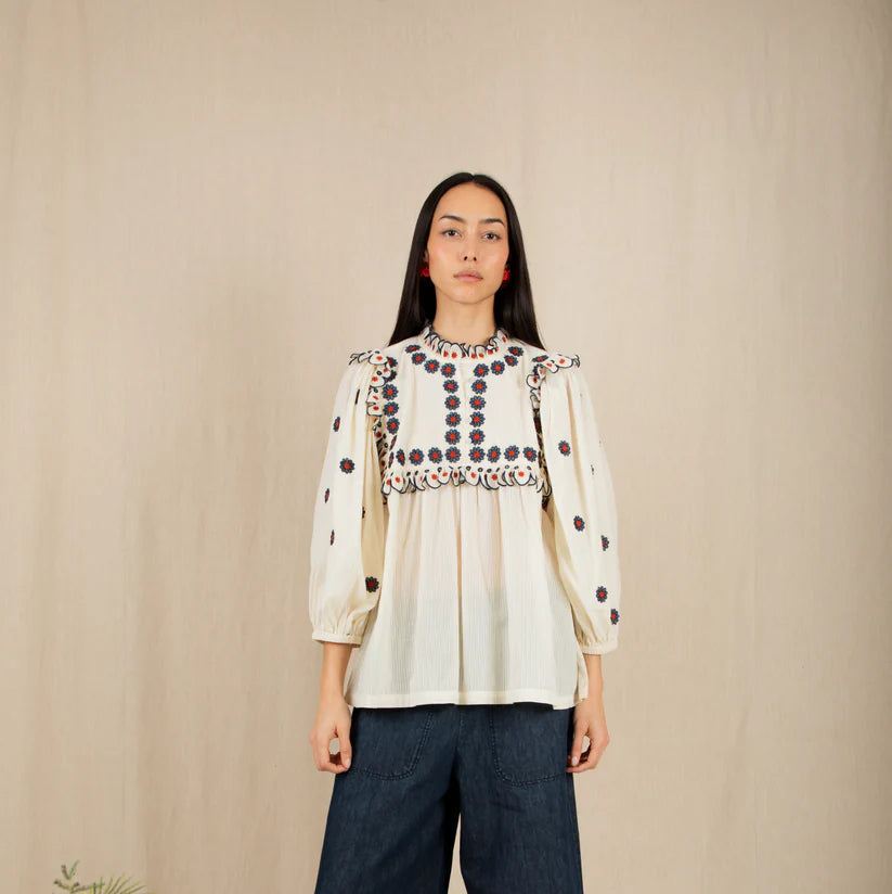 Woman wearing a patterned blouse and dark pants standing against a beige wall with yellow flowers on the left.