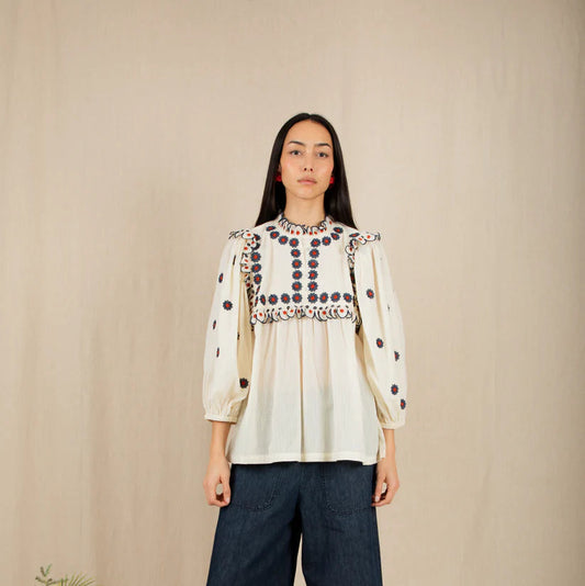 Woman wearing a patterned blouse and dark pants standing against a beige wall with yellow flowers on the left.