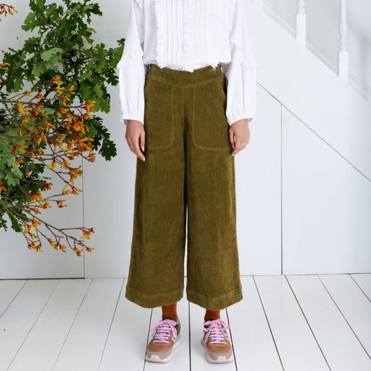 Person wearing a white blouse and brown corduroy pants standing in a minimalistic room with a plant.
