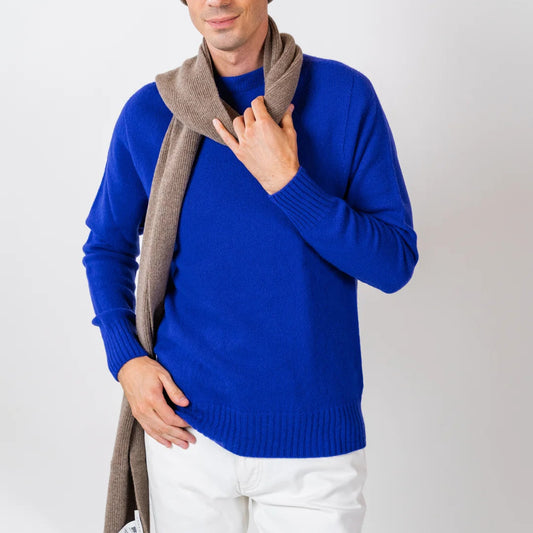 A man wearing a bright royal blue knit sweater, a taupe-brown knit beanie, and a matching taupe-brown knit scarf, paired with white trousers, photographed against a light gray studio background.